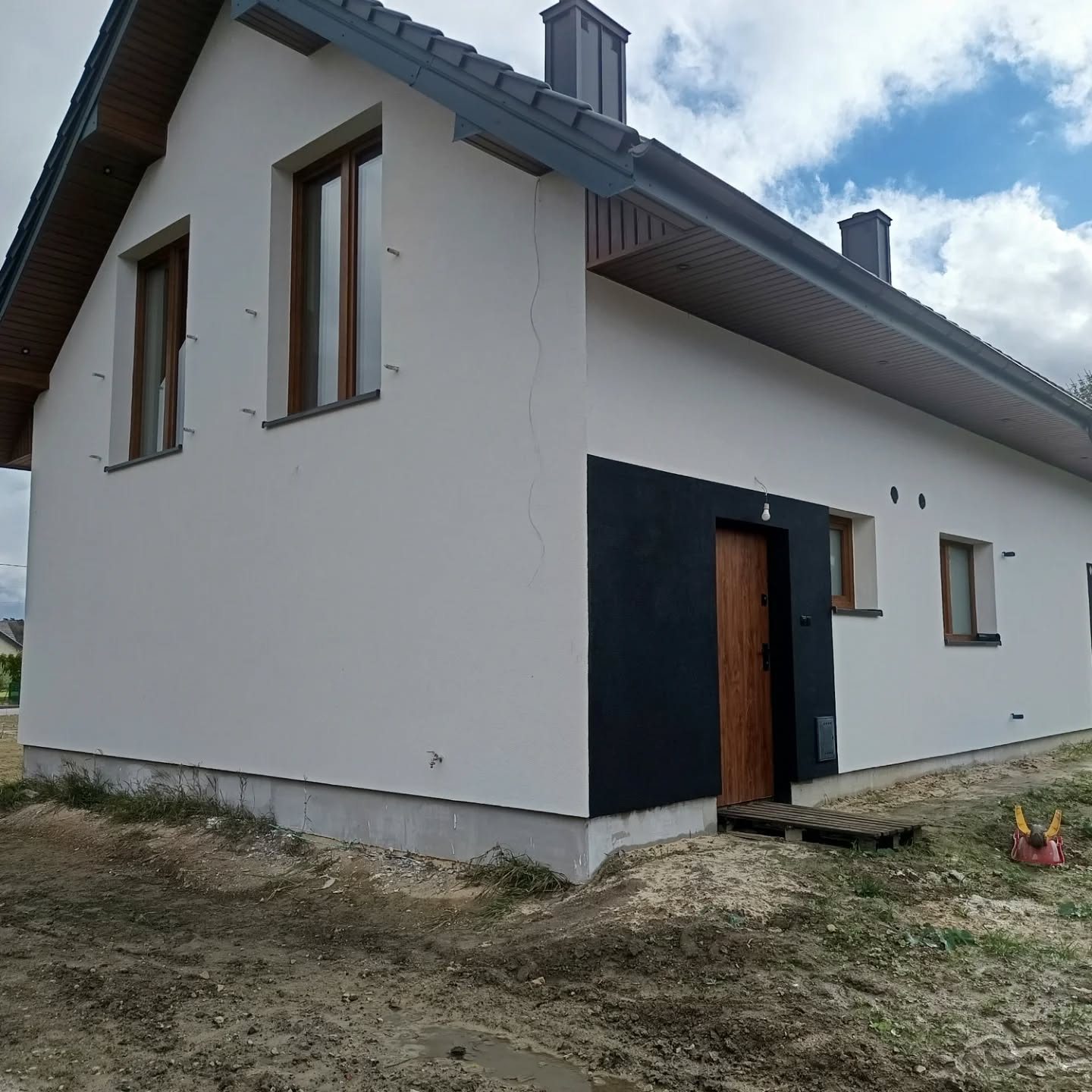 Jasna elewacja domu z czarnym wejściem, drewnianymi oknami i brązową podbitką